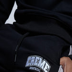 Load image into Gallery viewer, A close-up of a black sweatsuit featuring an embroidered CREME MONTREAL logo in white and blue on the pants. The hand rests casually in the pocket, highlighting the sleek design of the clothing.
