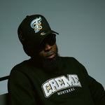 Load image into Gallery viewer, A person wearing a black cap with a blue and gold embroidered logo, sunglasses, and a black sweatshirt featuring the bold CREME MONTREAL logo is seated in a chair. The clothing exudes a stylish, urban vibe.
