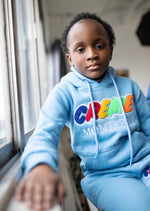 Load image into Gallery viewer, A child wearing a light blue hoodie with colorful chenille text sits by a window, gazing forward. The comfortable clothing features the word Montreal in embroidered letters, adding a playful touch.
