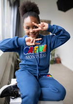 Load image into Gallery viewer, A child wearing a blue sweatsuit with colorful chenille patch embroidery sits on a windowsill, smiling and making peace signs with both hands. The cozy outfit showcases stylish kids clothing.
